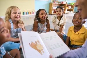 Female Primary Or Elementary School Teacher Reads Story To Multi-Cultural Class Seated In Classroom
